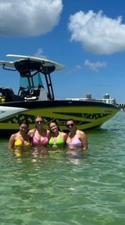 Four friends in colorful swimsuits standing waist-deep in clear turquoise water beside a yellow-and-white motorboat with a bridge and sunny blue sky in the background — summer boating day on a coastal inlet.