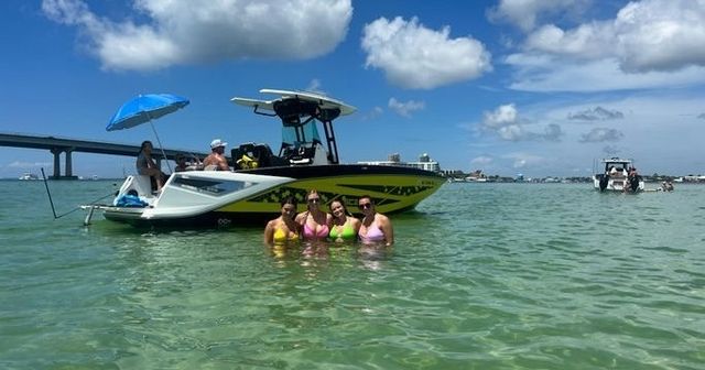 Four friends in colorful swimsuits standing waist-deep in clear turquoise water beside a yellow-and-white motorboat with a bridge and sunny blue sky in the background — summer boating day on a coastal inlet.