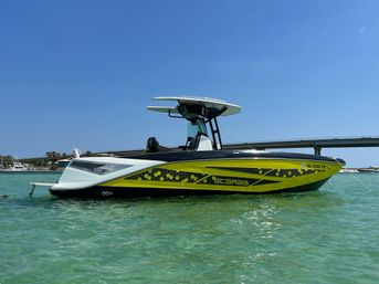 Sleek yellow-green sport powerboat with a T-top anchored in crystal-clear turquoise coastal water near a low bridge under a bright blue sky