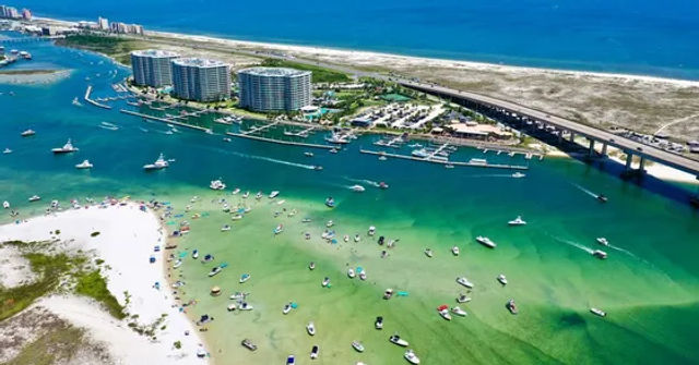 Aerial view of a turquoise coastal inlet and sandy barrier island with dozens of anchored boats, a marina, a curved highway bridge and beachfront condominium towers on a sunny day.