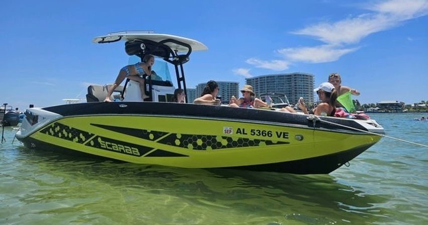 Yellow-and-black powerboat anchored in clear shallow coastal water with a group of friends lounging under a sunny blue sky and waterfront condos in the background