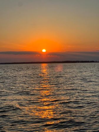 Golden sunset over open water with the sun sinking behind a low coastline, warm orange sky and shimmering reflection stretching across rippling sea.
