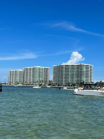 Sunny waterfront scene of curved condominium towers along a bay, with motorboats bobbing in green-blue water under a clear blue sky.