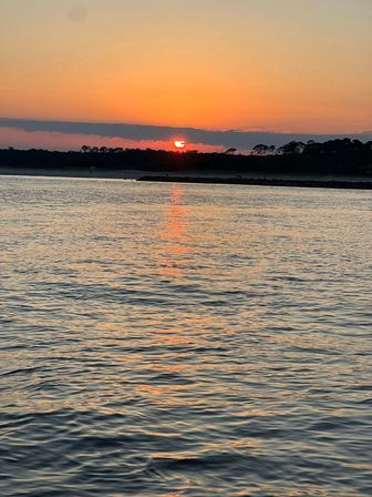 Vibrant orange sunset over a tree-lined coast, sun dipping behind clouds with a shimmering reflection across rippled bay waters.