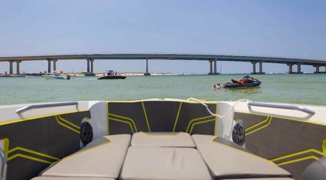 View from a boat bow toward a long coastal bridge over turquoise bay, with powerboats, a jet ski and people enjoying a sunny day.