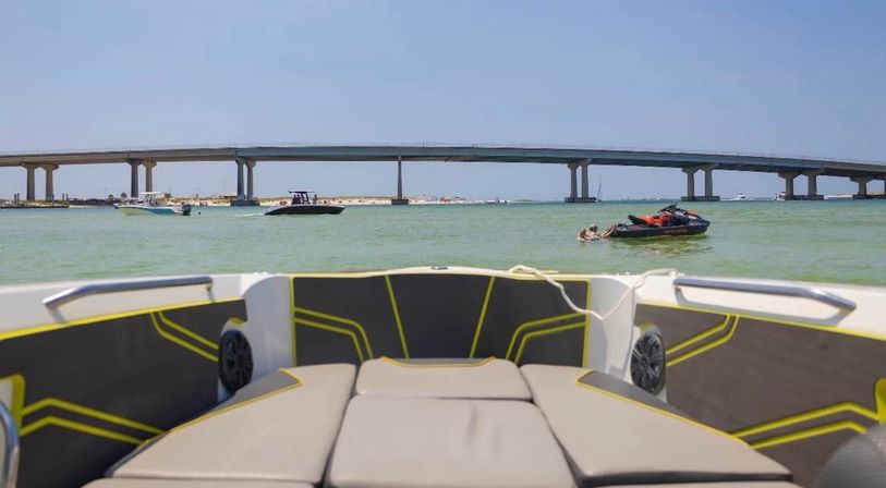 View from a boat bow toward a long low coastal bridge with recreational boats and a jet ski on turquoise water under a clear summer sky