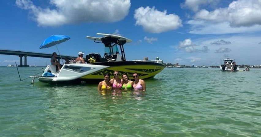Four friends in colorful bikinis wading in clear shallow bay water beside a black-and-yellow center-console boat with a blue umbrella, bridge and coastal skyline under a sunny blue sky