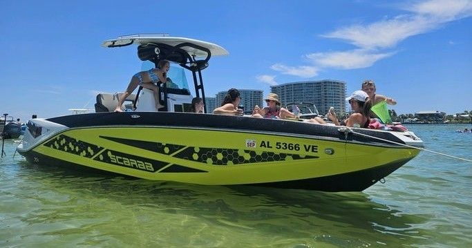 Lime-green and black powerboat anchored in shallow clear water near beachfront condominiums, people in swimsuits relaxing and enjoying a sunny blue-sky day on board.