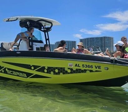 Bright yellow-and-black recreational speedboat anchored in clear shallow water, people relaxing under a T-top on a sunny day with beachfront condos visible in the background.