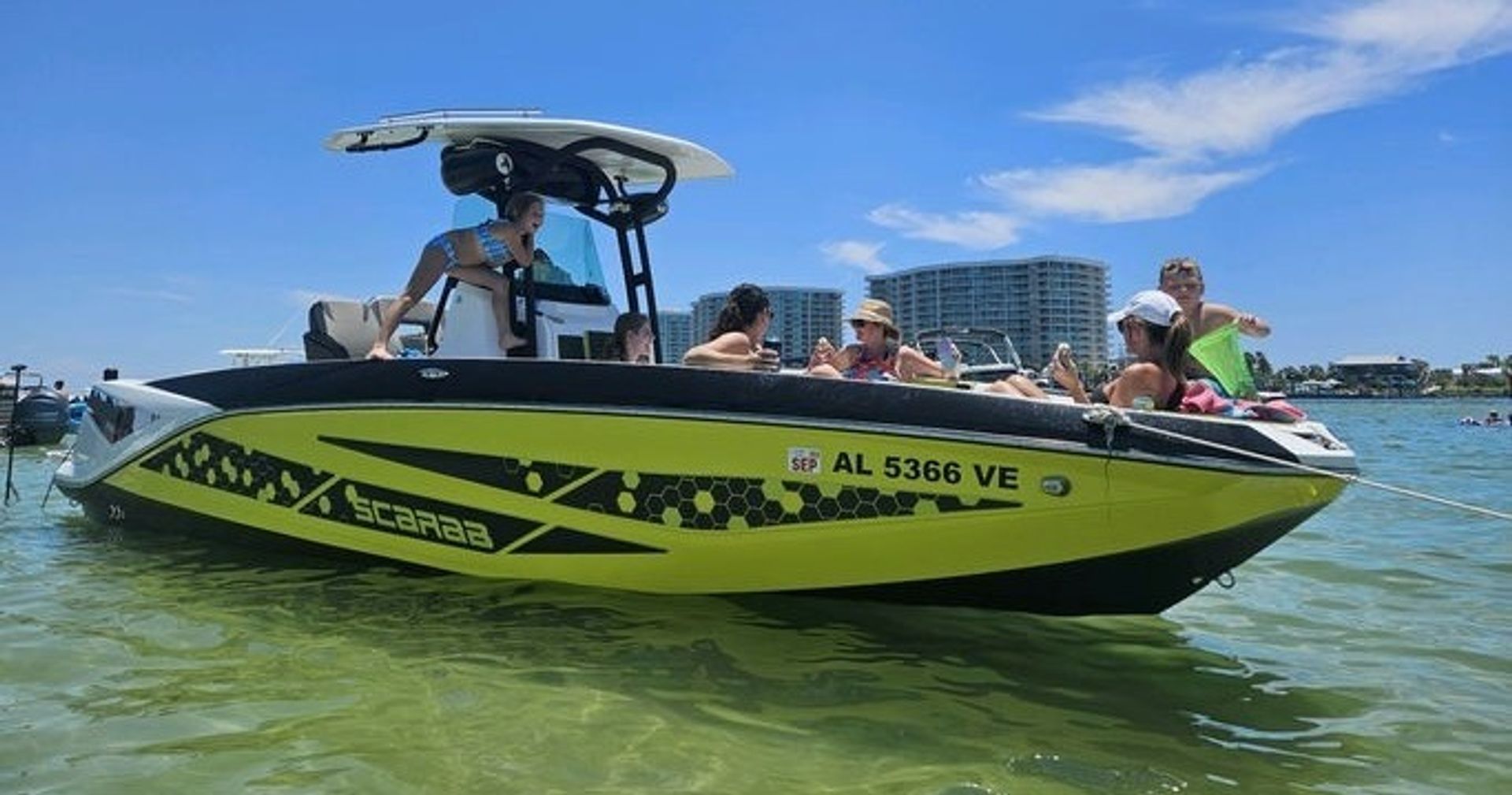 Bright yellow-and-black recreational speedboat anchored in clear shallow water, people relaxing under a T-top on a sunny day with beachfront condos visible in the background.