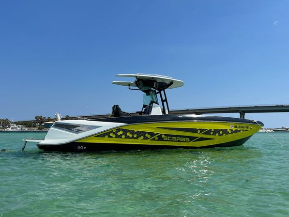 Chartreuse and black speedboat with T-top moored in clear turquoise bay under a bright blue sky, with a bridge and shoreline in the background.