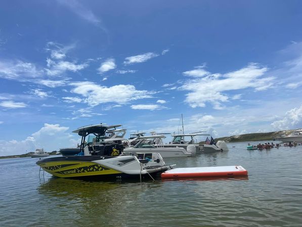 Sunny coastal sandbar scene with a yellow-and-black powerboat and other anchored boats, an orange inflatable swim platform, and a group of people floating nearby under a bright blue sky.