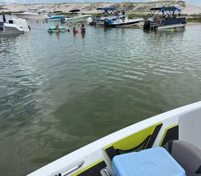 From a boat bow, light-blue cooler and cushioned seats in foreground; calm bay with anchored pontoon and powerboats near a sandy sandbar where people swim and float on tubes under a cloudy summer sky.