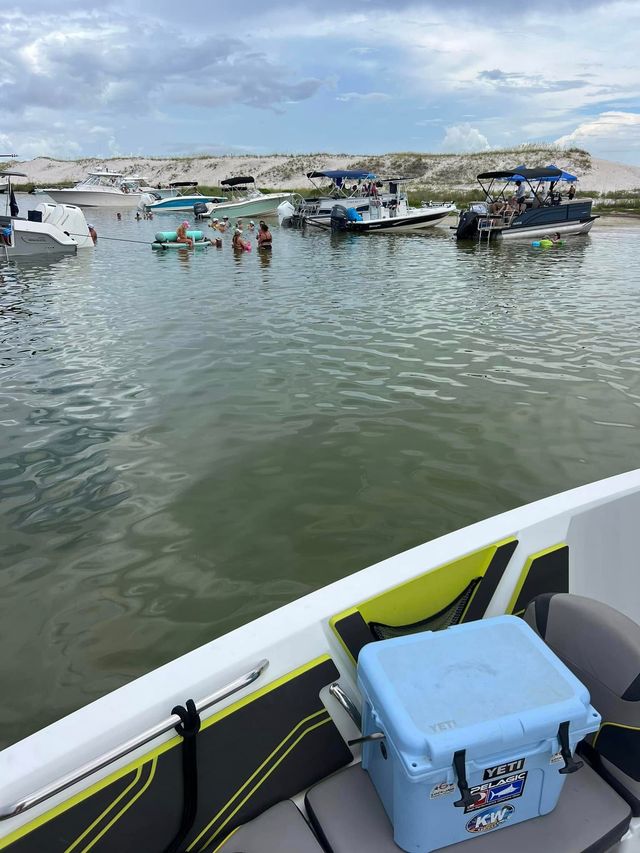 From a boat bow, light-blue cooler and cushioned seats in foreground; calm bay with anchored pontoon and powerboats near a sandy sandbar where people swim and float on tubes under a cloudy summer sky.