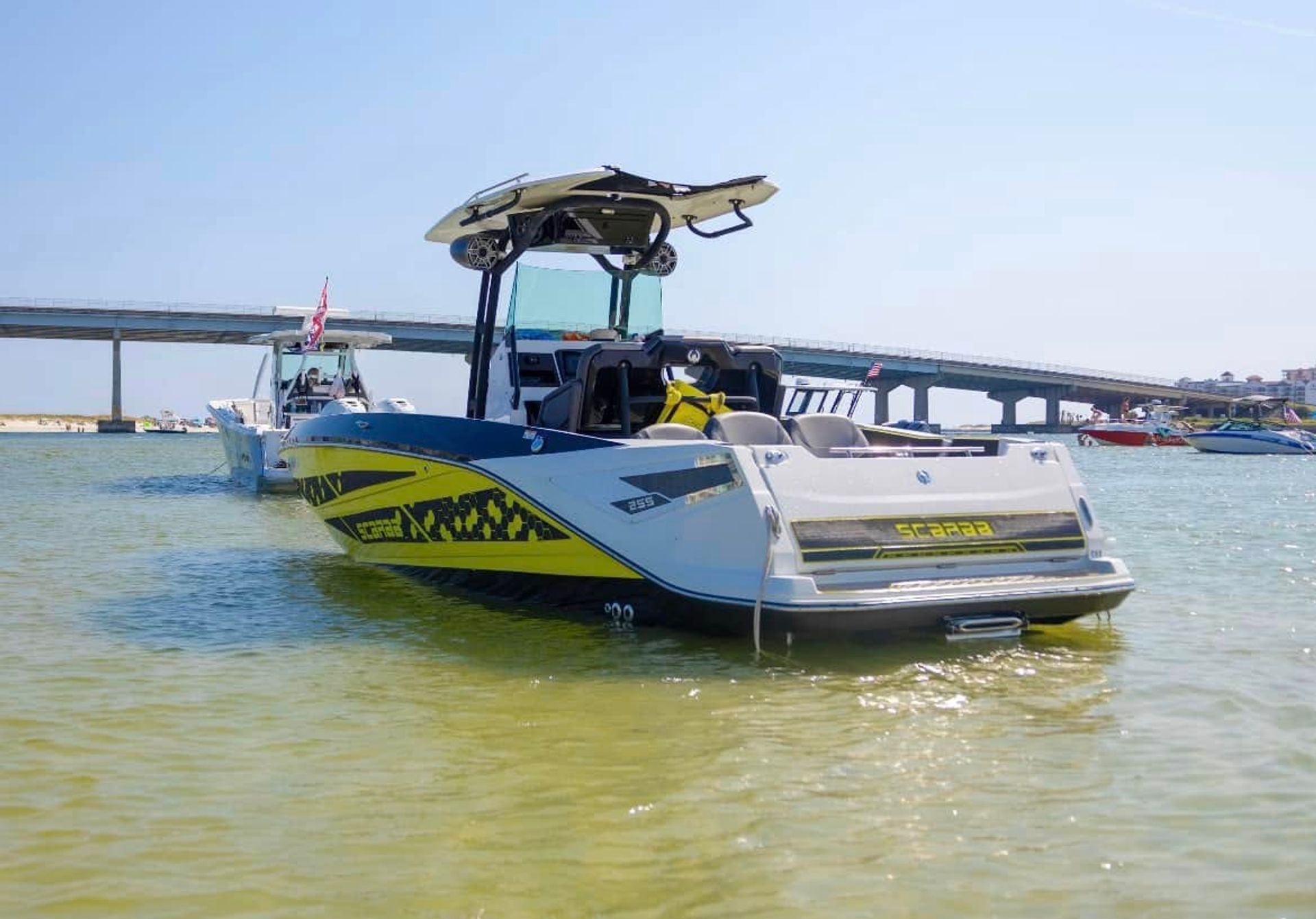 Bright yellow-and-white speedboat with a roof-mounted board anchored in shallow clear green water near a coastal bridge on a sunny day, with other boats and a sandy shoreline in the background.