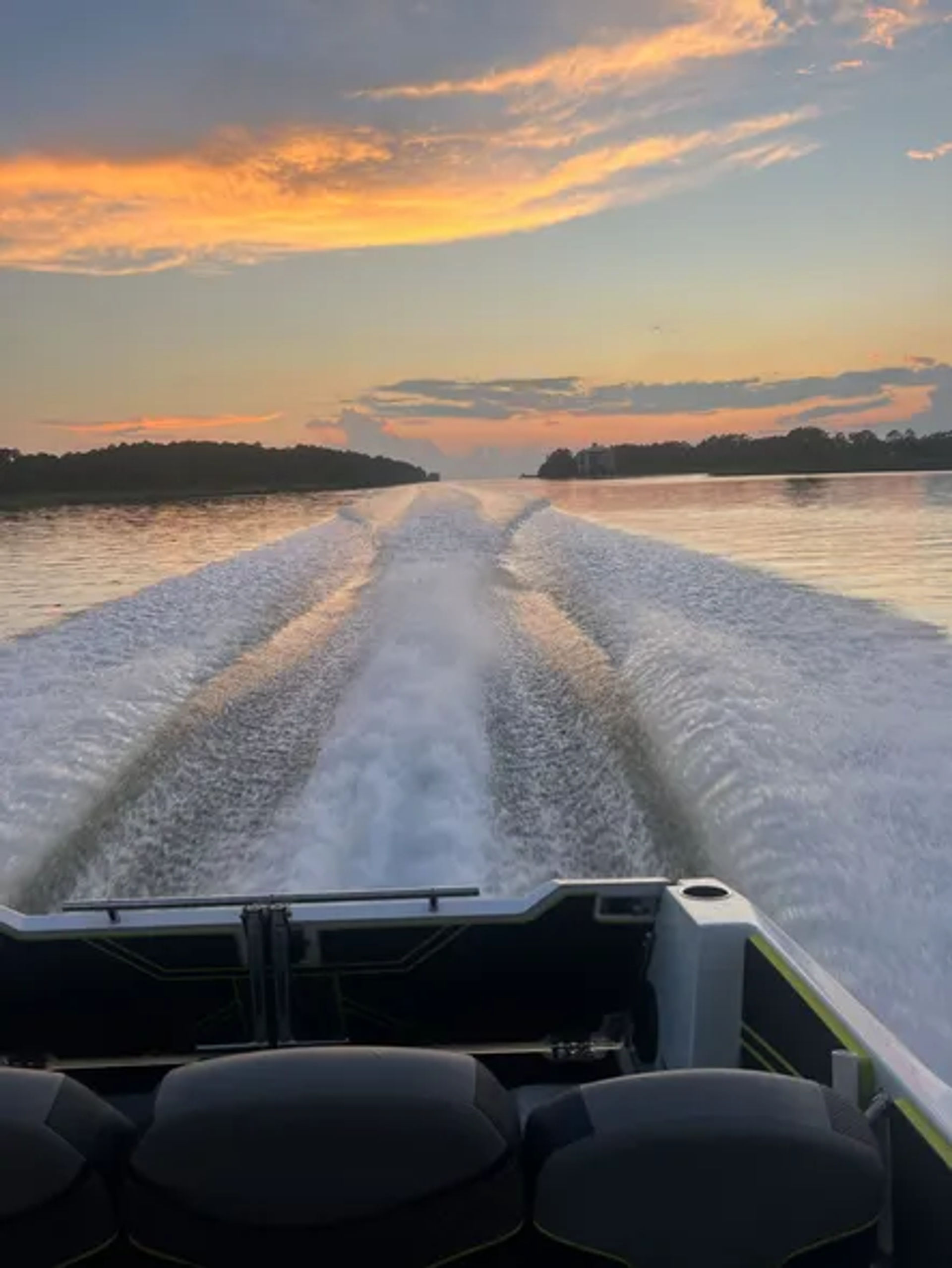 View from a motorboat's stern at sunset — twin wake trails streak across a calm lake toward a tree-lined horizon under glowing orange-pink clouds.