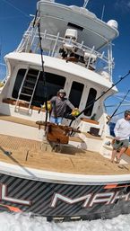 Offshore sportfishing yacht cockpit with teak deck, two anglers at a fighting chair handling gold big-game reels and multiple outriggers, blue sky and open ocean visible