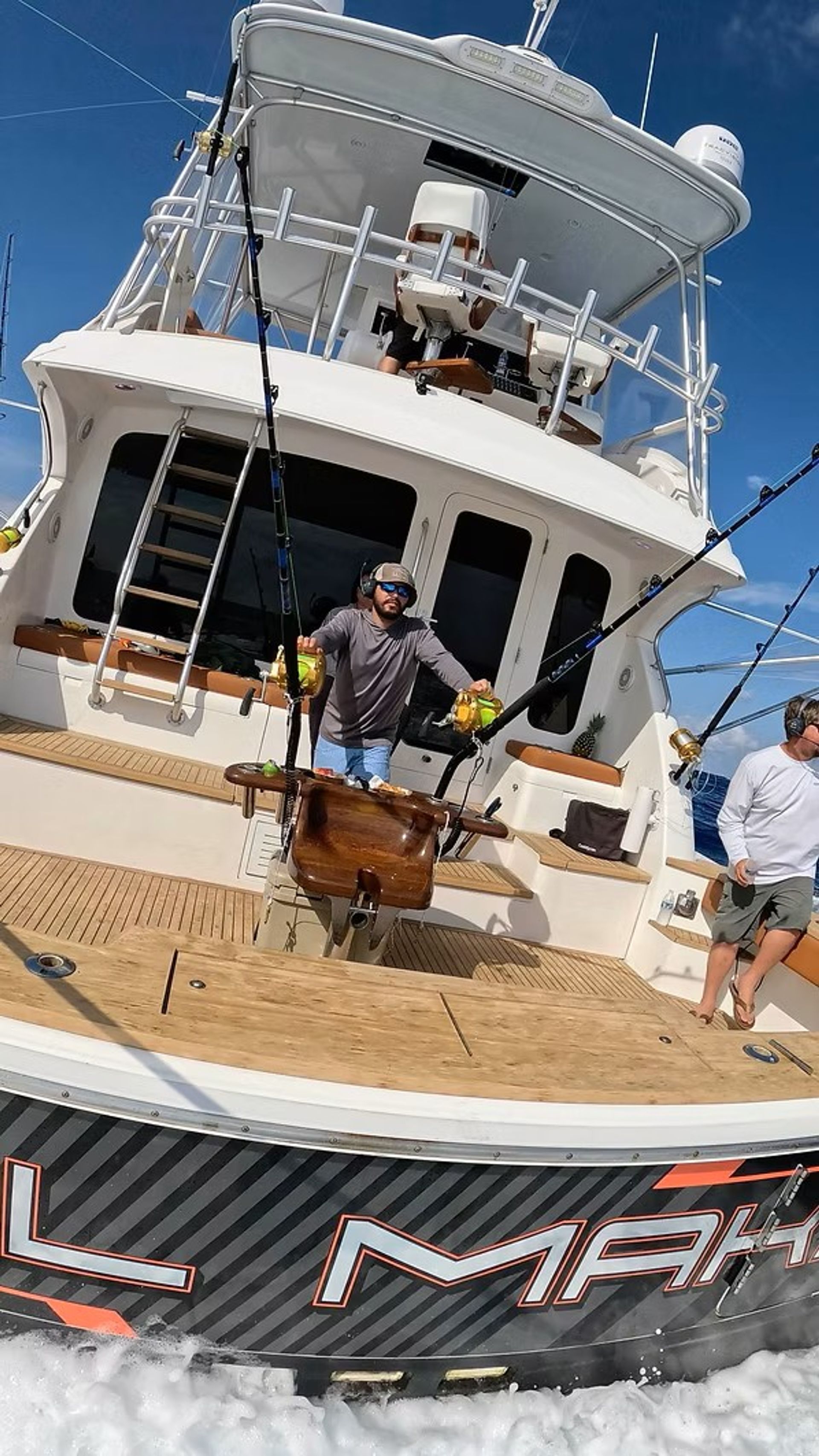 Offshore sportfishing yacht cockpit with teak deck, two anglers at a fighting chair handling gold big-game reels and multiple outriggers, blue sky and open ocean visible