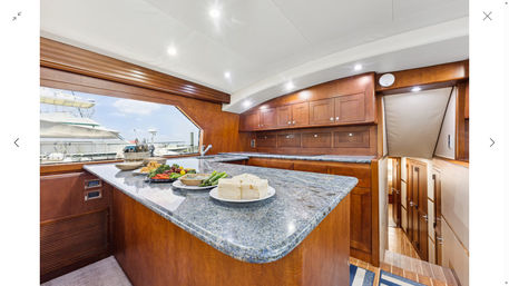 Luxury yacht galley with polished granite island topped with plates of cheese, vegetables and appetizers, rich wood cabinetry, recessed lighting and a window view of a marina with docked boats.