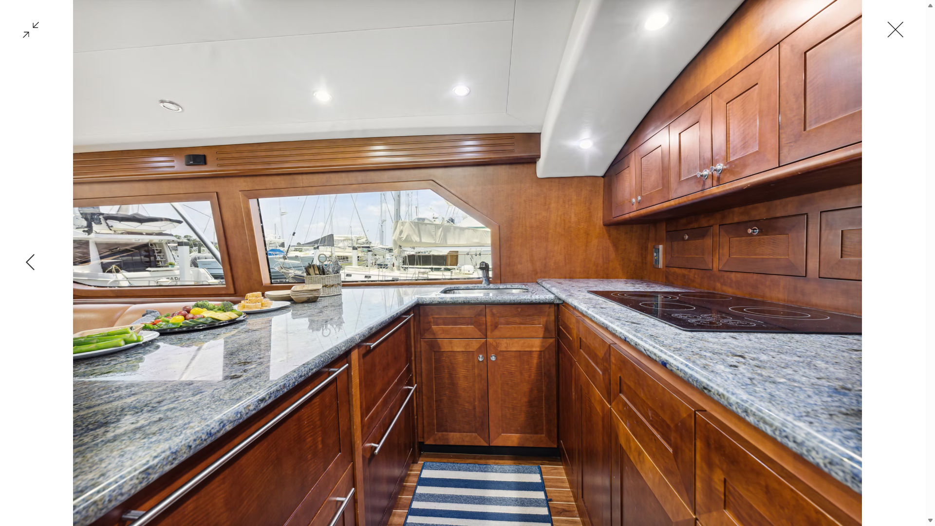 Sunlit luxury yacht galley with polished cherry wood cabinets, blue-gray granite countertops, induction cooktop, stainless sink, striped runner and marina view through large windows.
