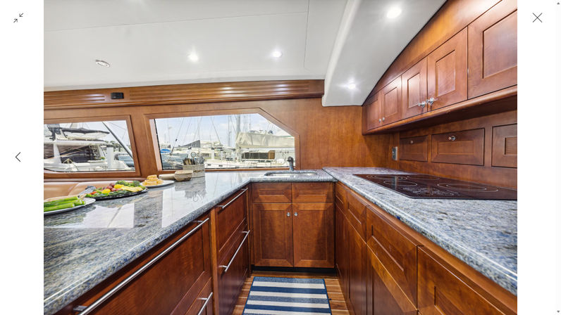 Sunlit luxury yacht galley with polished cherry wood cabinets, blue-gray granite countertops, induction cooktop, stainless sink, striped runner and marina view through large windows.