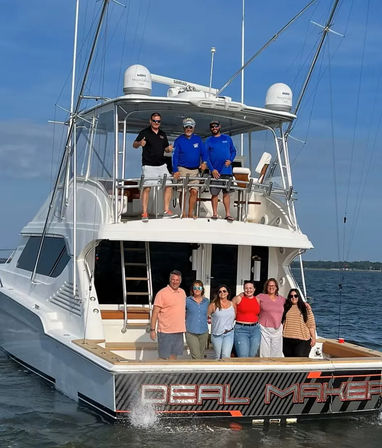Nine adults posing on a large white sportfishing yacht’s stern and flybridge; sunny blue sky, calm coastal waters, radar domes and fishing outriggers visible.
