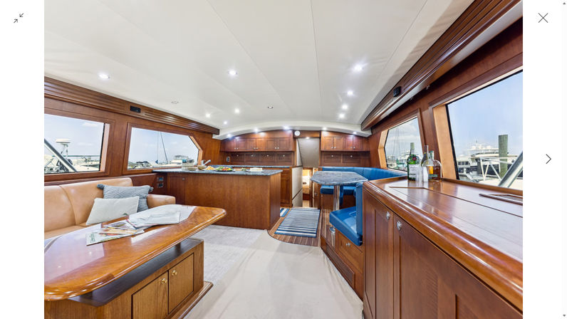 Bright luxury yacht interior salon at the marina featuring polished mahogany cabinetry, leather sofa, blue upholstered dinette, galley island, panoramic windows and recessed lighting