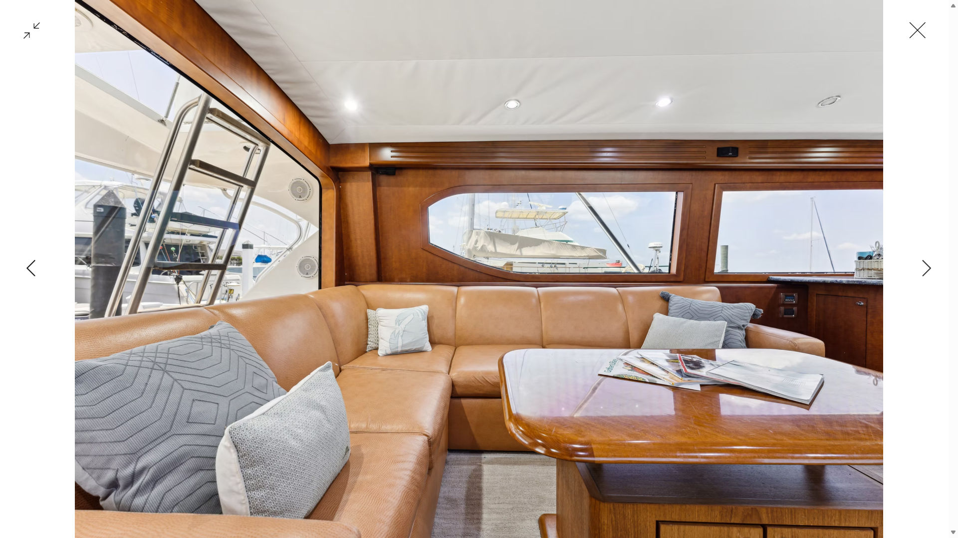 Sunlit luxury yacht salon with tan leather L-shaped sofa, textured gray pillows, glossy wood table with magazines, warm wood paneling and large windows framing a marina with boats.