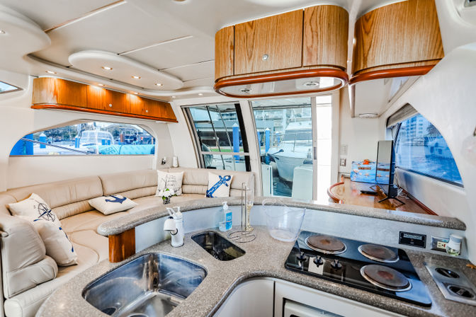 Sunlit luxury yacht interior galley and salon with curved leather seating, nautical pillows, wood cabinetry, granite countertop with stainless sink and electric cooktop, sliding glass doors opening to a marina view