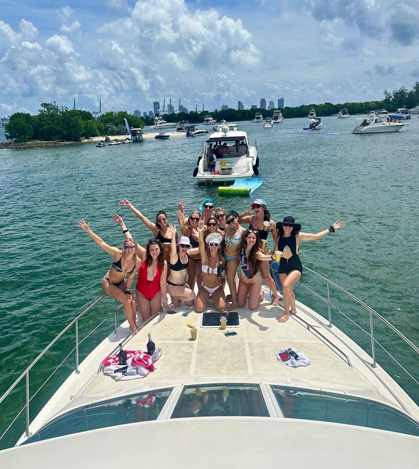 Lively group of friends in swimsuits posing on the bow of a yacht during a sunny boat party in a tropical bay with other anchored boats and a distant city skyline