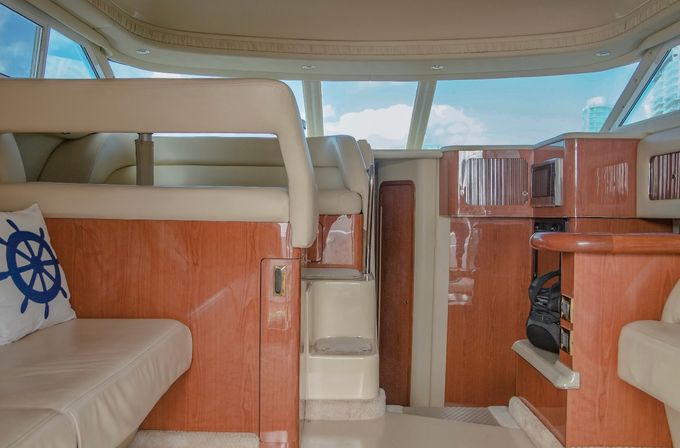 Sunlit luxury yacht interior salon with cream leather seating, nautical wheel throw pillow, glossy cherry-wood cabinetry and steps up to the helm through large windows showing blue sky and distant coastal skyline.