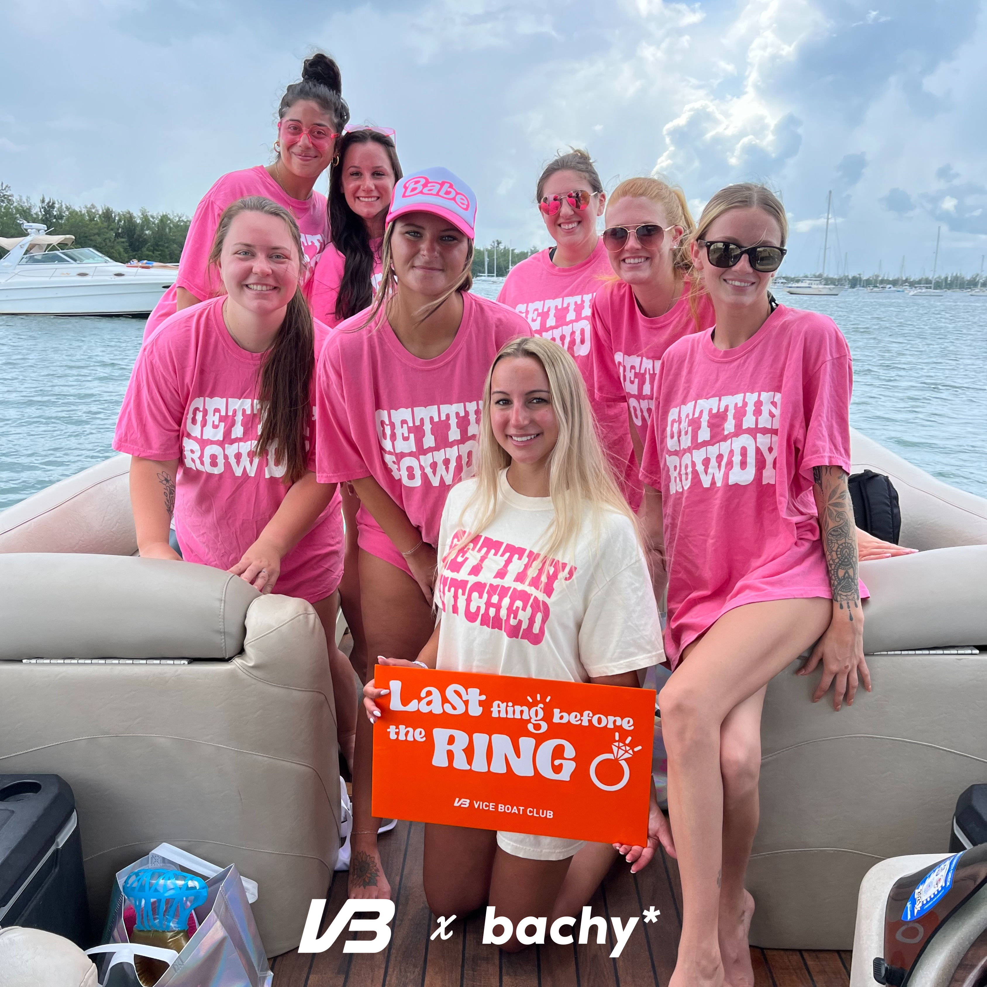 Bachelorette boat party at a marina: eight smiling women on a pontoon, most in matching pink 'Gettin' Rowdy' shirts and sunglasses, holding an orange 'Last fling before the ring' sign under a cloudy sky.