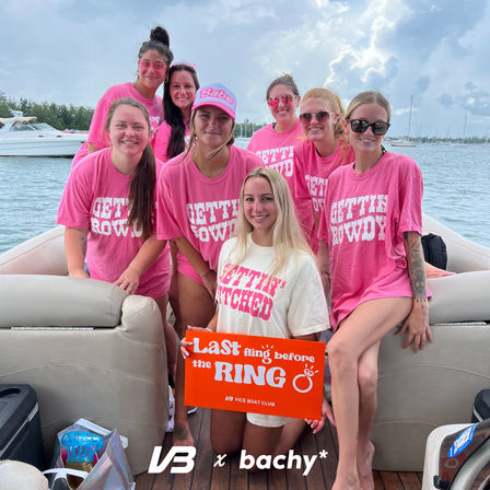 Bachelorette boat party at a marina: eight smiling women on a pontoon, most in matching pink 'Gettin' Rowdy' shirts and sunglasses, holding an orange 'Last fling before the ring' sign under a cloudy sky.