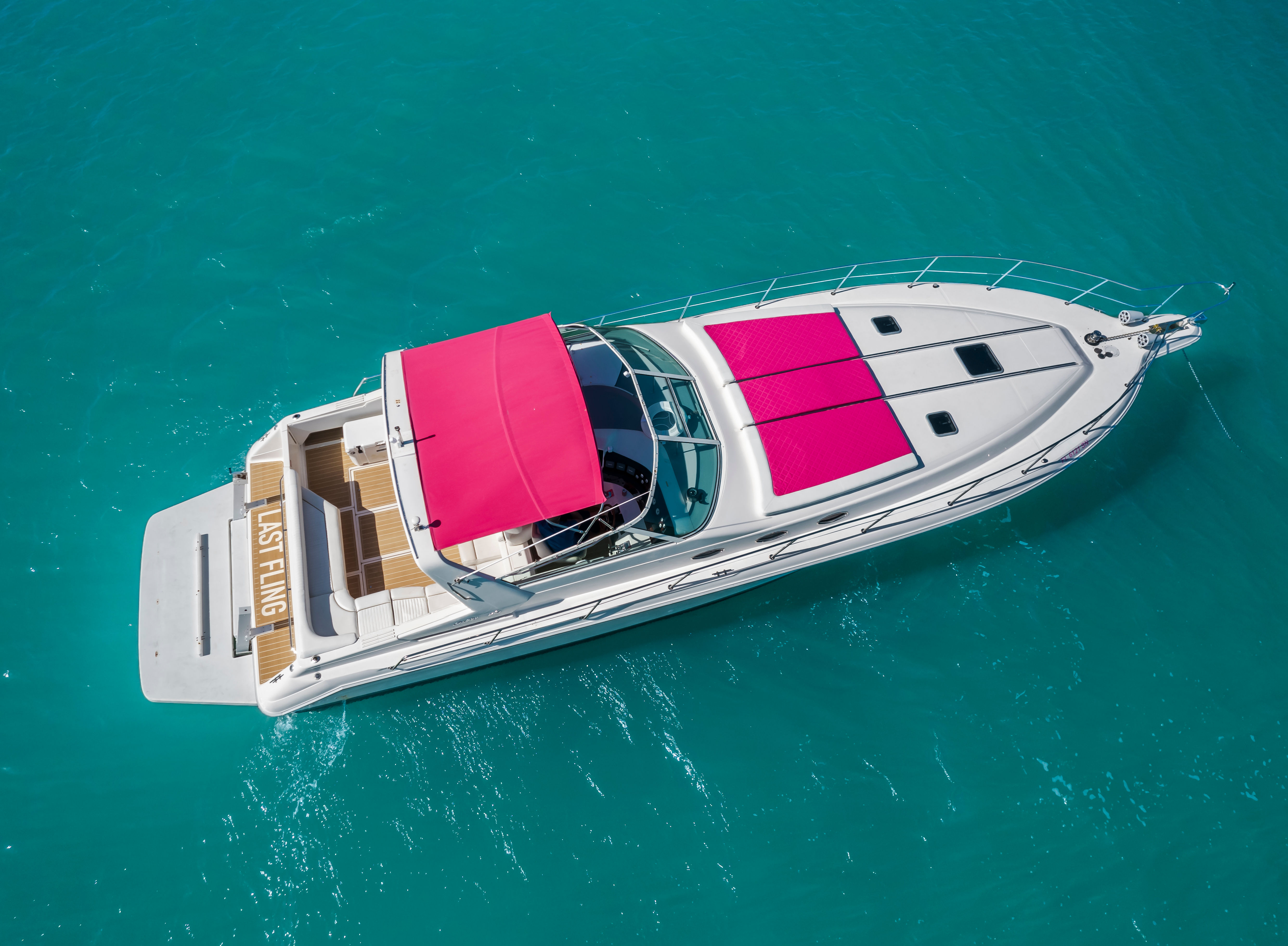 Aerial view of a white luxury motor yacht with a bright pink canopy and matching pink sun pads floating on clear turquoise water.