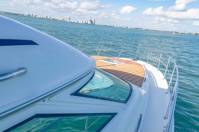 Bow of a white yacht with teak foredeck and stainless railing gliding through turquoise coastal waters, distant city skyline on the horizon under a sunny blue sky