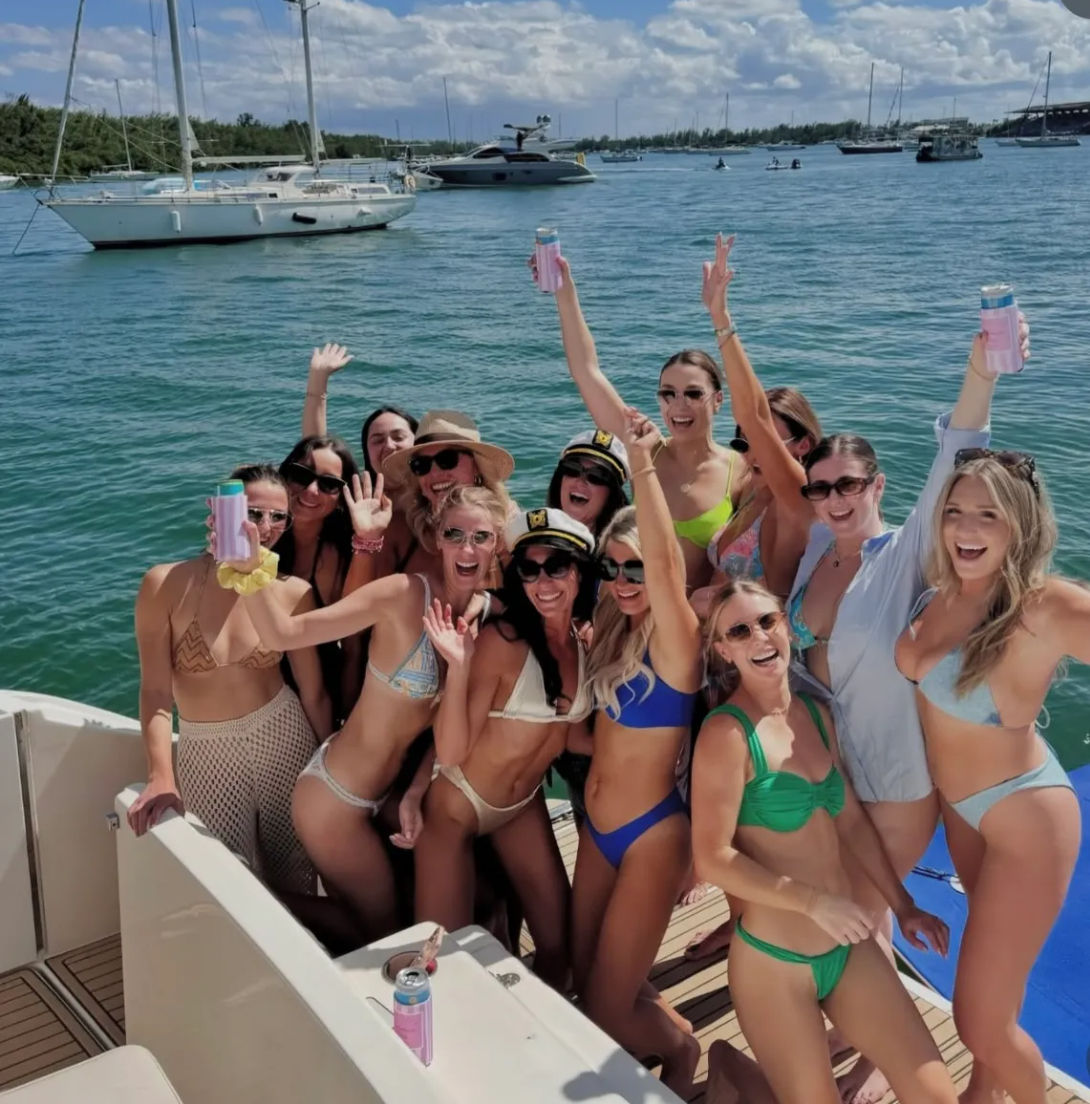 Group of friends in colorful bikinis cheering with drinks on a yacht over turquoise water, sailboats and a sunny marina in the background
