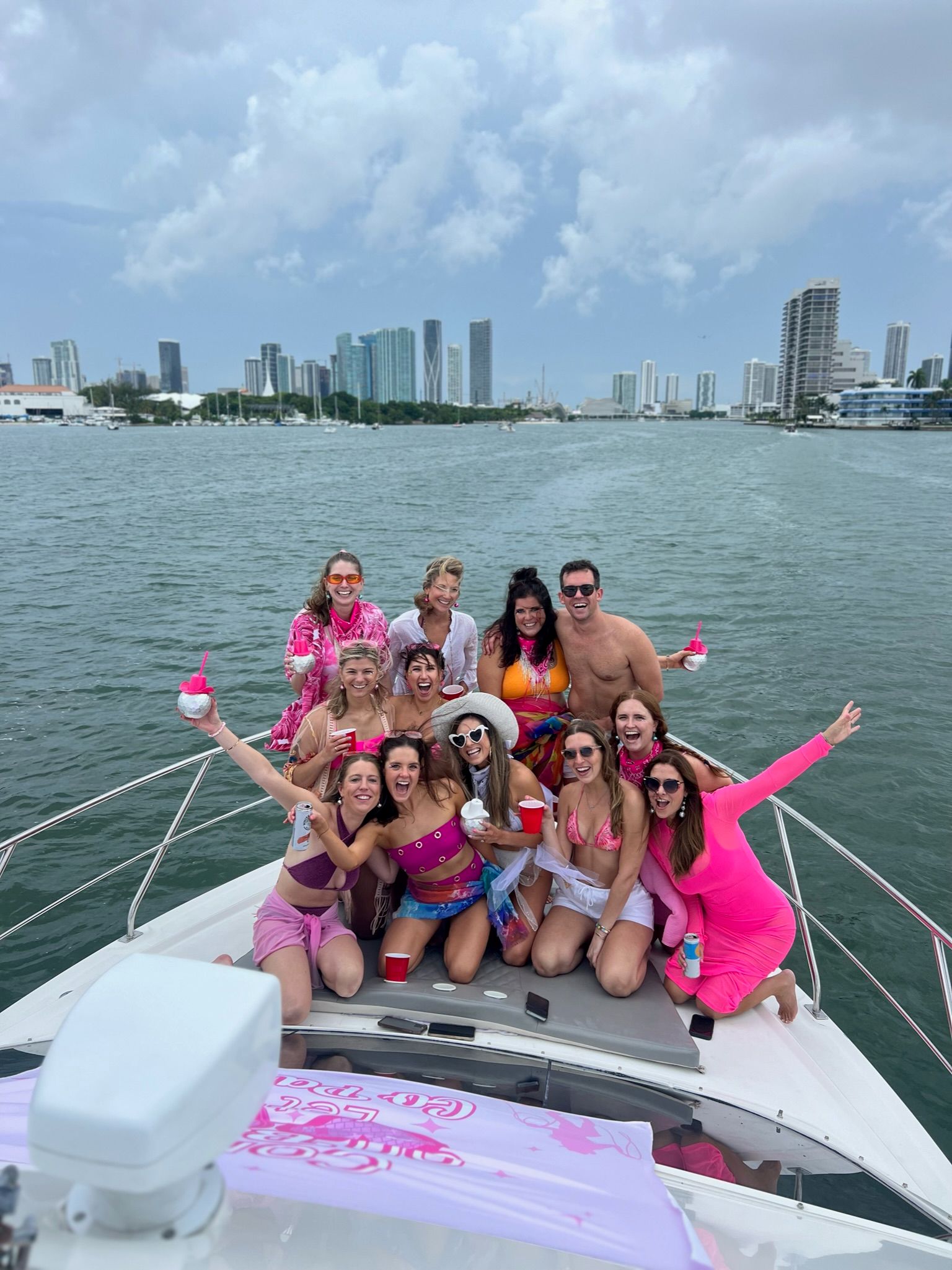 Group of friends in bright pink swimsuits and party clothes celebrating with drinks on a yacht bow in Biscayne Bay, Miami skyline and cloudy sky in the background