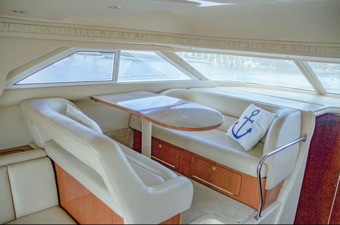 Sunlit luxury yacht cabin dining area with white leather banquette, wooden table, built-in storage drawers, anchor-printed pillow and panoramic waterfront view through large side windows