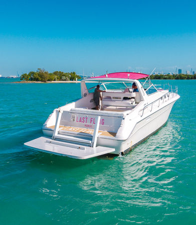 White motor yacht with pink canopy cruising turquoise Miami waters near a small island and distant city skyline on a sunny day.