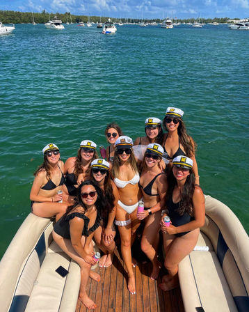 Group of friends in swimsuits wearing captain hats on a sunny party boat in turquoise bay with anchored yachts and a tree-lined shore