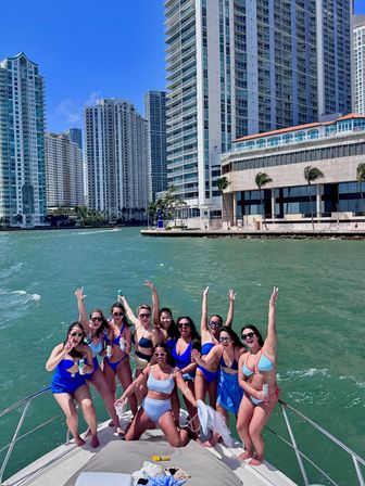 Group of women in colorful swimsuits cheering on a yacht off the sunny Miami waterfront, turquoise water with high-rise condo skyline in the background.