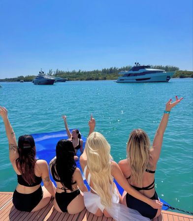 Four friends in swimsuits sit on a yacht platform with arms raised, overlooking turquoise water and anchored luxury yachts under a clear sunny sky in a tropical bay