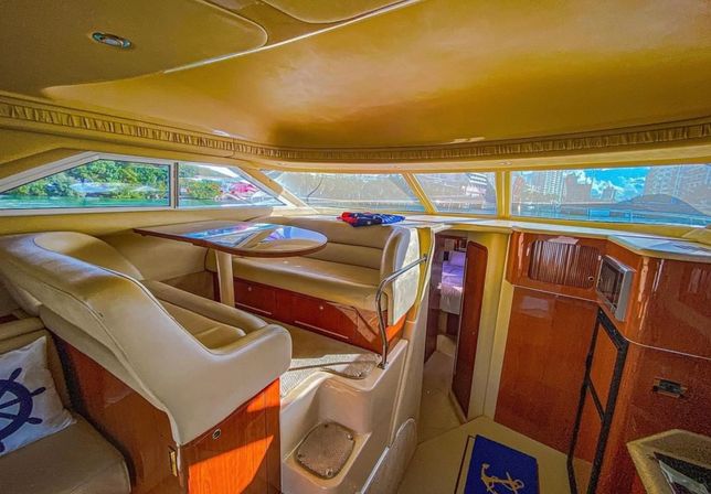 Sunlit yacht interior featuring a cozy beige leather dinette with oval wood table, glossy cherry wood cabinetry, steps to a lower cabin and panoramic windows revealing a marina and city skyline view.