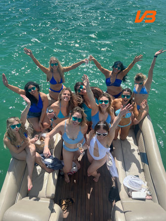 Aerial view of a lively summer boat party: a group of women in blue and white swimsuits and sunglasses cheering and holding champagne on the deck of a pontoon boat over sparkling emerald-green water.