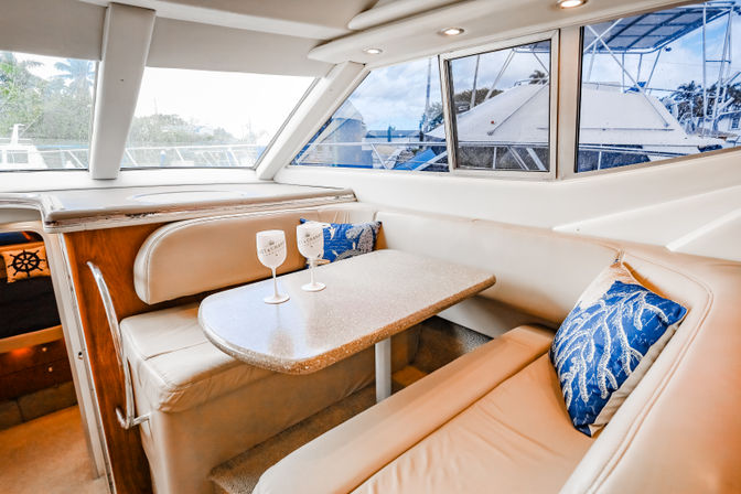 Cozy yacht dinette with beige leather seating, speckled table topped with two wine glasses, blue coral-pattern throw pillows and large windows overlooking a marina with nearby boats.