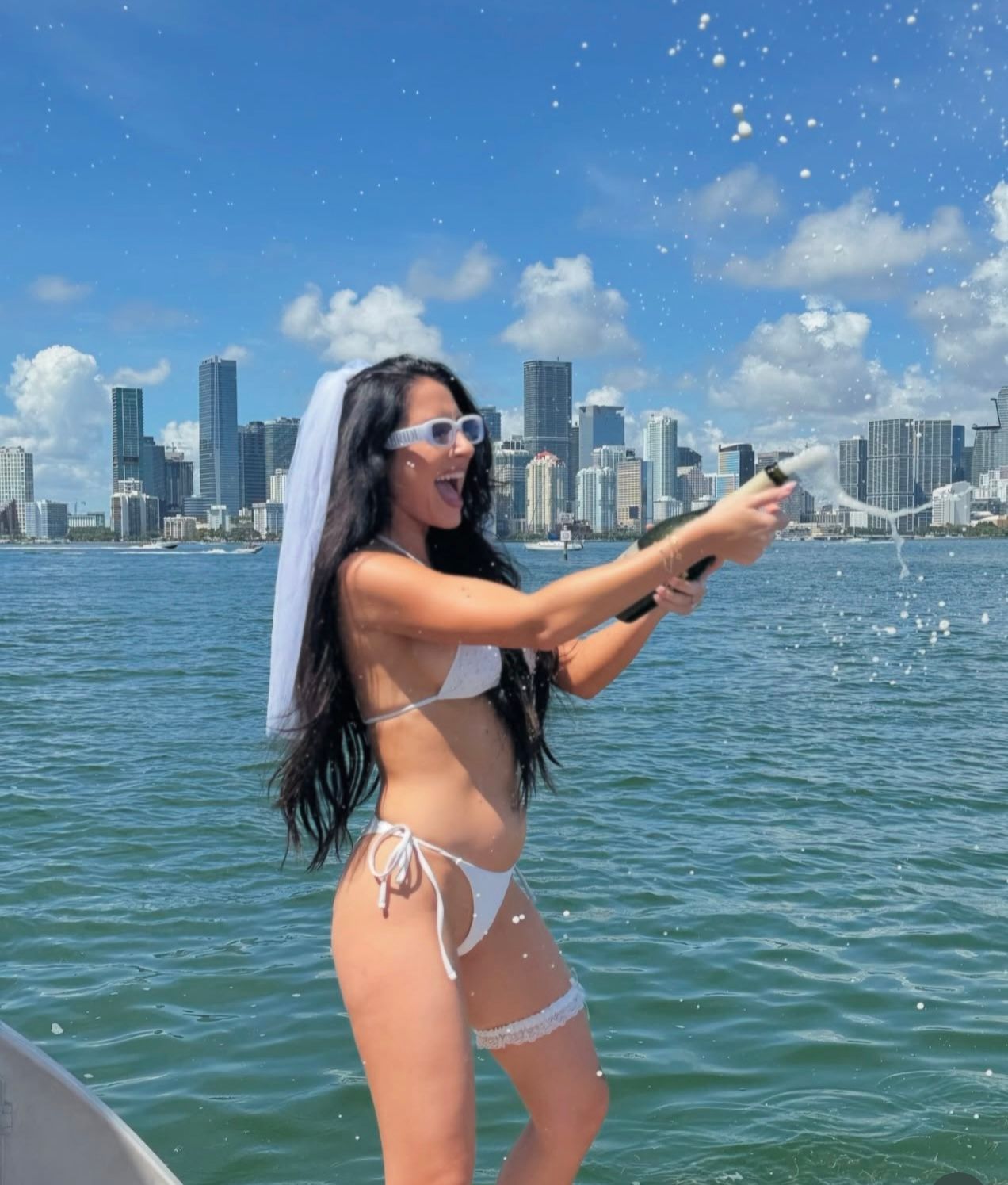 Bachelorette in a white bikini and veil wearing sunglasses pops champagne on a boat, champagne spray flying against a blue-sky coastal city skyline over calm bay water — festive summer waterfront celebration.