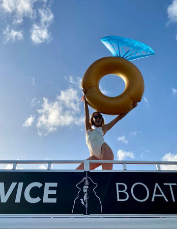 Person in a white swimsuit on a boat deck holding a giant gold inflatable ring with a blue "gem" aloft against a bright blue sky — fun summer boat party vibe.
