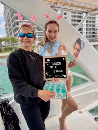 Two friends on a boat at a marina celebrating a bachelorette — woman with pink novelty headband and blue feather boa holding a 'BRIDE TO B' letterboard, friend in sunglasses serving a tray of blue shots with waterfront condos in the background.