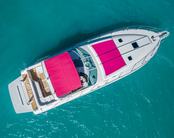 Aerial overhead view of a white luxury yacht with bright-pink canopy and matching sun pads, sun deck and swim platform visible as it floats on clear turquoise water.
