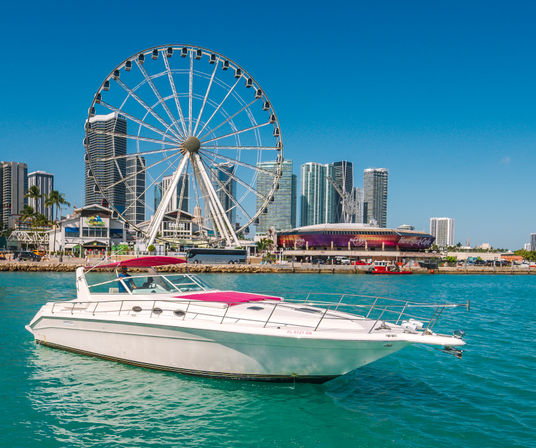 White motor yacht on turquoise harbor waters with a large waterfront Ferris wheel and modern high-rise skyline under a bright blue sky